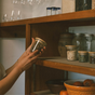 Hands hold a glass jar with Me Mother Earth Solid Bamboo Mason Jar Lids, reaching toward a wooden pantry shelf filled with assorted food jars and a bowl of oranges.