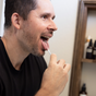 A man in a black shirt uses Me Mother Earth's Stainless Steel Tongue Scrapers + Travel Tins to clean his tongue in a bathroom, with shelves and bottles visible, highlighting an oral care routine.