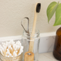 A bamboo toothbrush, Me Mother Earth Stainless Steel Tongue Scrapers + Travel Tins, and nail clippers sit in a glass jar by cotton swabs and a green leafy plant, creating an inviting oral care setup on the bathroom counter.
