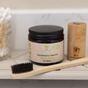 A glass jar of Me Mother Earth Toothpaste Tablets with Fluoride - Zero Waste sits on a bathroom shelf next to a bamboo toothbrush, wooden holder, and cotton swabs. The label reads Peppermint, 65 tablets—a perfect travel-friendly solution.