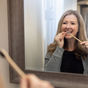 A woman with long brown hair smiles while flossing in front of a bathroom mirror, wearing a gray sweater. Me Mother Earth Toothpaste Tablets with Fluoride – Zero Waste sit near a toothbrush in the foreground.