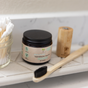 A jar of Me Mother Earth Toothpaste Tablets in an amber glass jar (fluoride-free), a bamboo toothbrush with black bristles, a bamboo container, and a glass jar of cotton swabs are arranged on a white bathroom shelf.