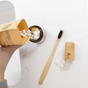 A hand pours Me Mother Earth Toothpaste Tablets 3 Month Refill Pouch - Fluoride-Free into a glass jar. A bamboo toothbrush and wooden travel case sit nearby on a white countertop with a few loose tablets.