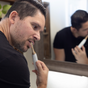 A man with short dark hair and a trimmed beard brushes his teeth in front of a bathroom mirror, wearing a black t-shirt and using Me Mother Earth Toothpaste Tablets 3 Month Refill Pouch - Fluoride-Free.