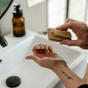 A person uses the Me Mother Earth Wood Nail Brush to clean their fingernails over a white sink. A forearm tattoo is visible, and a brown soap dispenser rests on the counter.