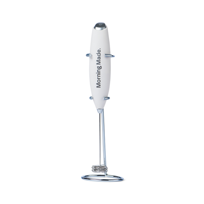 The Morning Made Modern Whisk is a handheld, battery-operated coffee frother with a white body, a chrome stand for upright storage, and a small electric whisk at the bottom.