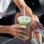 A person in shorts and a white shirt sits on black stairs in sunlight, holding a Morning Made Insulated Tumbler with a straw in one hand and white headphones in the other.