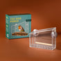 The Modern Sprout Window Bird Feeder, crafted from clear acrylic and featuring a wooden perch with sturdy suction cups, is displayed as a chic window accessory beside its green box adorned with an image of both the feeder and a bird—all arranged against a warm brown backdrop.