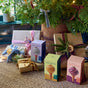 Gifts wrapped in brown and white paper with colorful ribbons and dried orange slices sit under a decorated tree. Three packages of Modern Sprout Watering Bulbs, Variety Pack, are displayed in front on a woven rug.