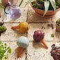 Three Modern Sprout Watering Bulbs from the Variety Pack in yellow, purple, and light blue with ribbed designs sit on a tiled surface among potted plants and a glass watering can, providing a stylish, easy solution for plant care.