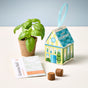 A small potted basil plant from the Modern Sprout Home Grown Ornaments collection sits beside a decorative house-shaped planter box, a Genovese basil seed packet, and two soil pellets on a white surface with a light blue background—making it the perfect herb growing kit and ornament gift for any kitchen windowsill.