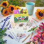 A Magical Minds Garden Kit by Modern Sprout sits on a white table alongside colored markers, flower seed packets, plant markers, sunflowers, snapdragons, and a blue cup—creating a vibrant, nature-inspired scene reminiscent of an enchanting fairy garden kit for kids.