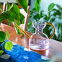 The Modern Sprout Handcrafted Borosilicate Glass Watering Can, with a yellow handle and spout, sits on a wooden table by green potted plants and a blue patterned runner. It also doubles as a stylish plant propagation vessel.