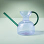 The Modern Sprout Handcrafted Borosilicate Glass Watering Can features a clear body, long green spout, and curved handle—perfect for watering or as a stylish plant propagation vessel. Shown on a pale surface with a light green background.