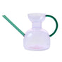 The Modern Sprout Handcrafted Borosilicate Glass Watering Can features a pinkish body, long left-angled green spout, and curved right green handle—ideal for watering or as a unique plant propagation vessel against a white background.