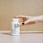 A bottle of Thorne Magnesium Glycinate is placed on a beige surface with a hand reaching for it. The label displays an abstract design and black text, highlighting this chelated magnesium supplement against a tan background.