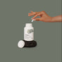 A hand holds a white capsule above an open Thorne Magnesium Glycinate bottle, set on dark textured stone with a plain green backdrop.