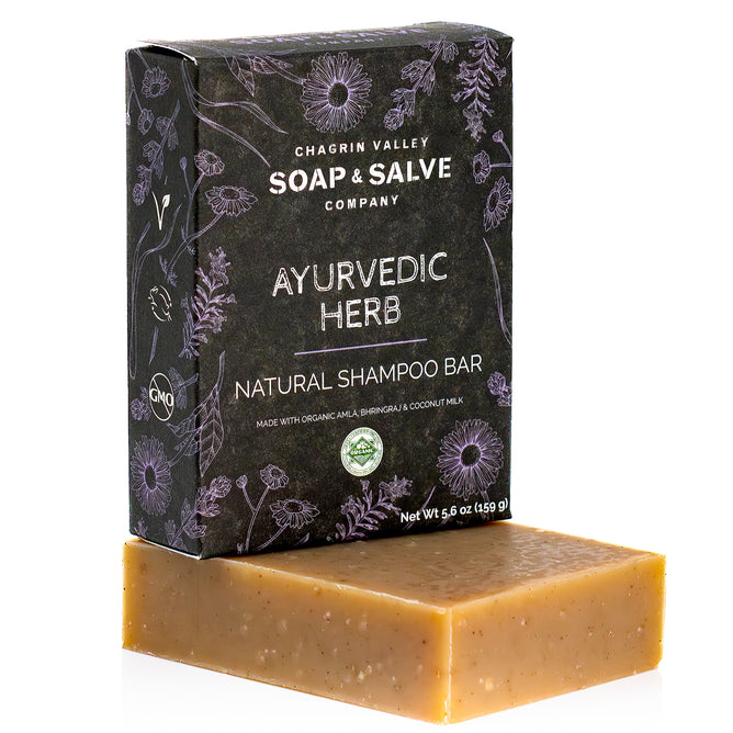 A brown rectangular Natural Shampoo Bar made with organic coconut milk rests in front of a Chagrin Valley Soap & Salve box labeled “Ayurvedic Herb Natural Shampoo Bar” with floral designs and product details.