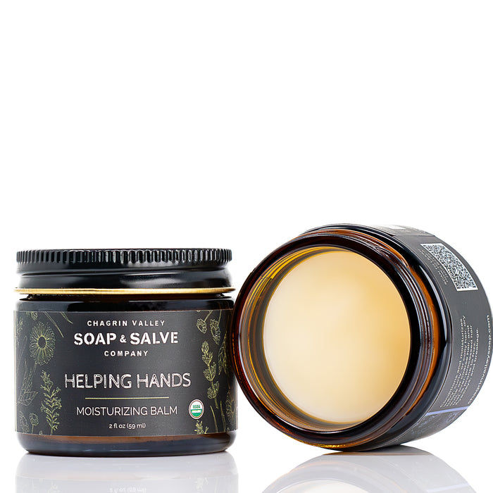 Two brown glass jars of Chagrin Valley Soap & Salve Hand Balm: Helping Hands are shown. One jar is open to reveal the organic balm inside, while the other is closed and displays the product label.