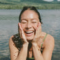 A woman smiles joyfully with her eyes closed, splashing Ursa Major’s Making Moves Milky Cleanser on her face for hydration while standing in a lake, surrounded by green hills and a blue sky.