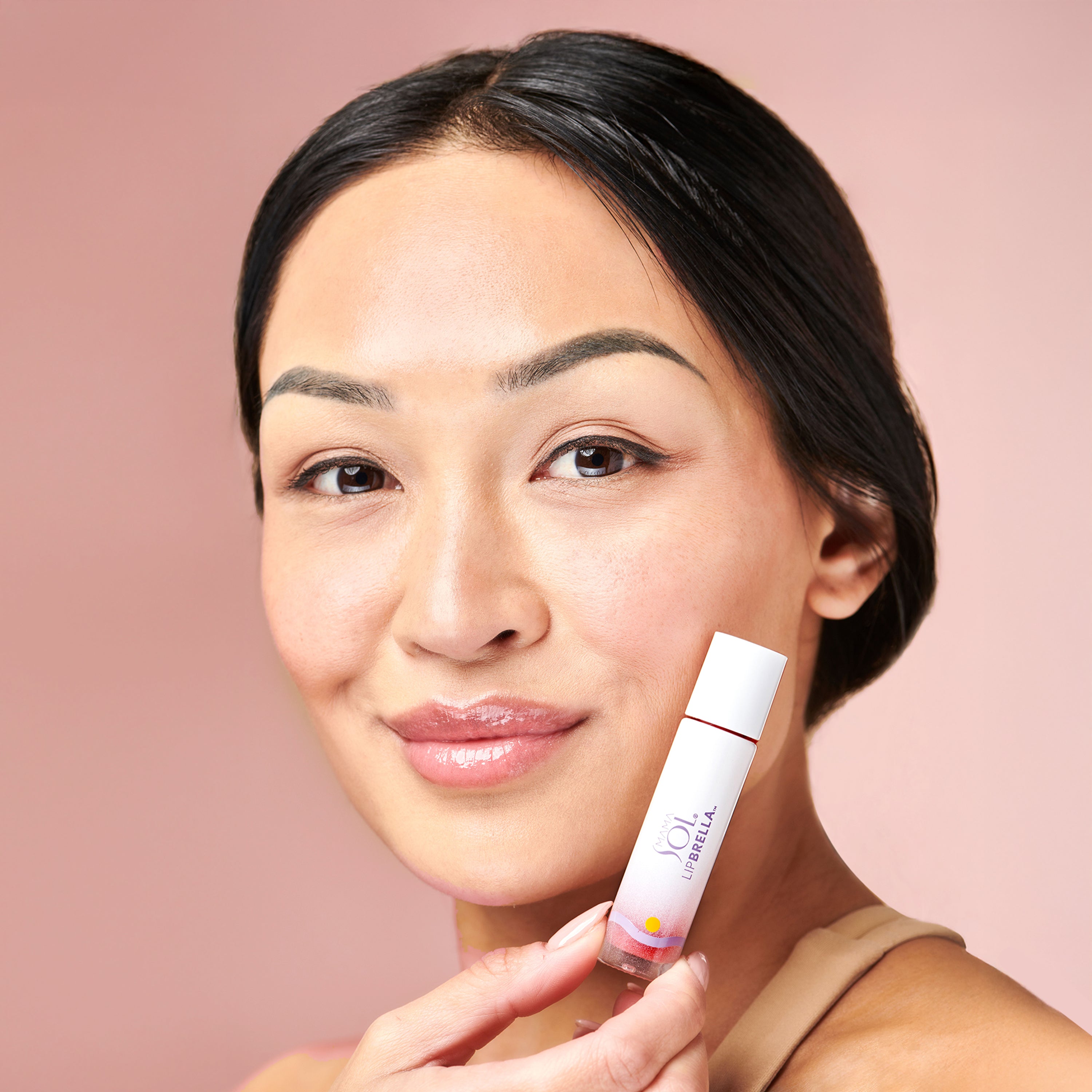 A woman with smooth, glowing skin holds Mama Sol’s LIPBRELLA 100% Mineral Tinted Lip Oil SPF 20 near her face, smiling gently against a soft pink background.