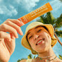 A smiling person in a yellow hat holds up an orange packet of blume’s SuperBelly Probiotic Hydration Powder - Sample, emphasizing its gut health benefits with probiotics, against a backdrop of palm trees and a vibrant blue sky.