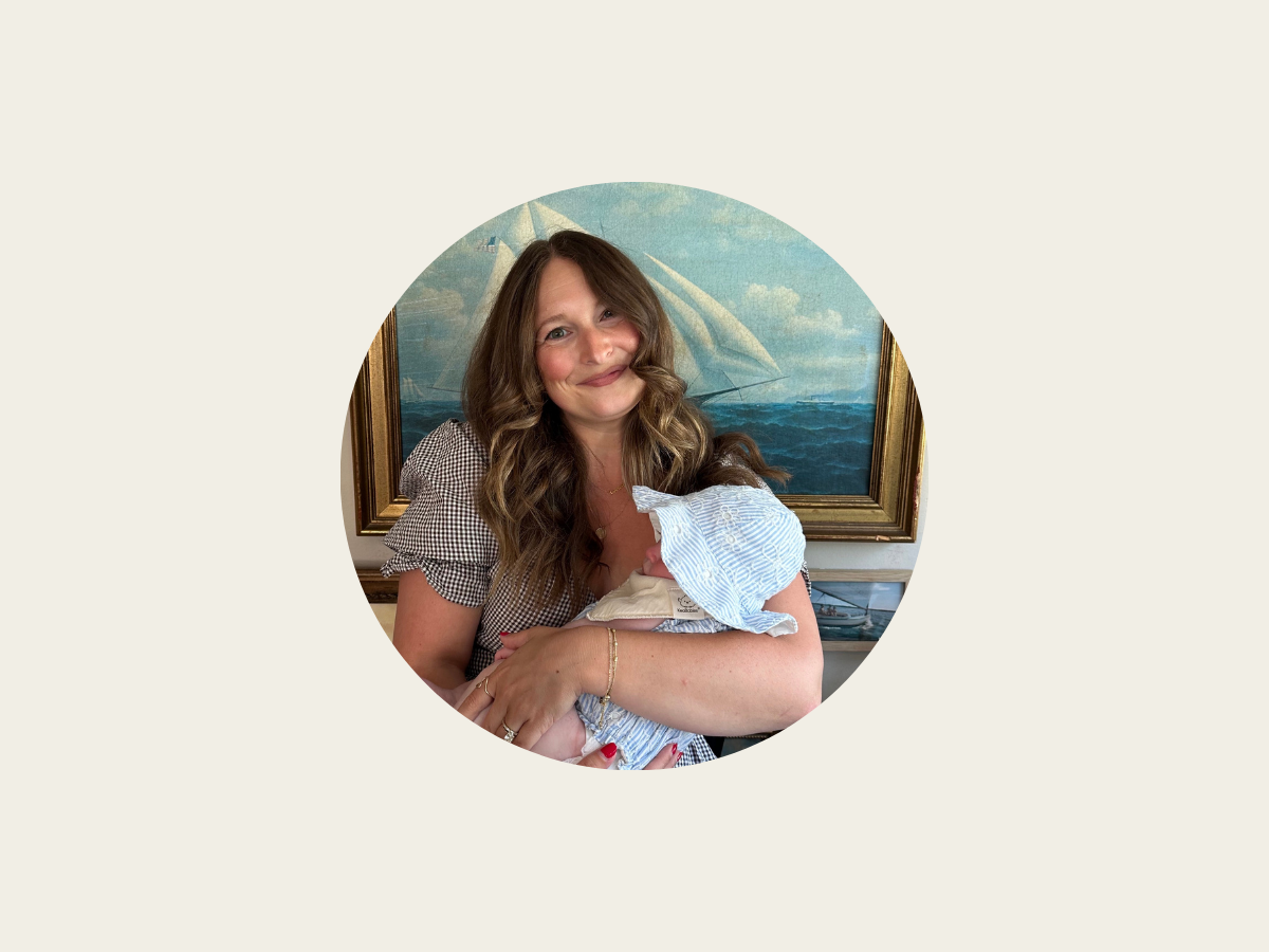 A woman with wavy brown hair smiles while holding a baby dressed in blue and white clothing. They are in front of a painting of a sailboat on the ocean, all set against a plain, light background.