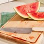 A Caraway Birch Wood Board holds a white-handled knife beside two watermelon slices. Set on a green textured mat, the FSC-certified board has juice droplets and stacked plates in the background.
