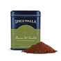 A tin labeled Spicewalla Deluxe Mexican Hot Chocolate sits beside a mound of rich dark cocoa powder, hinting at cayenne chili warmth, on a white background.