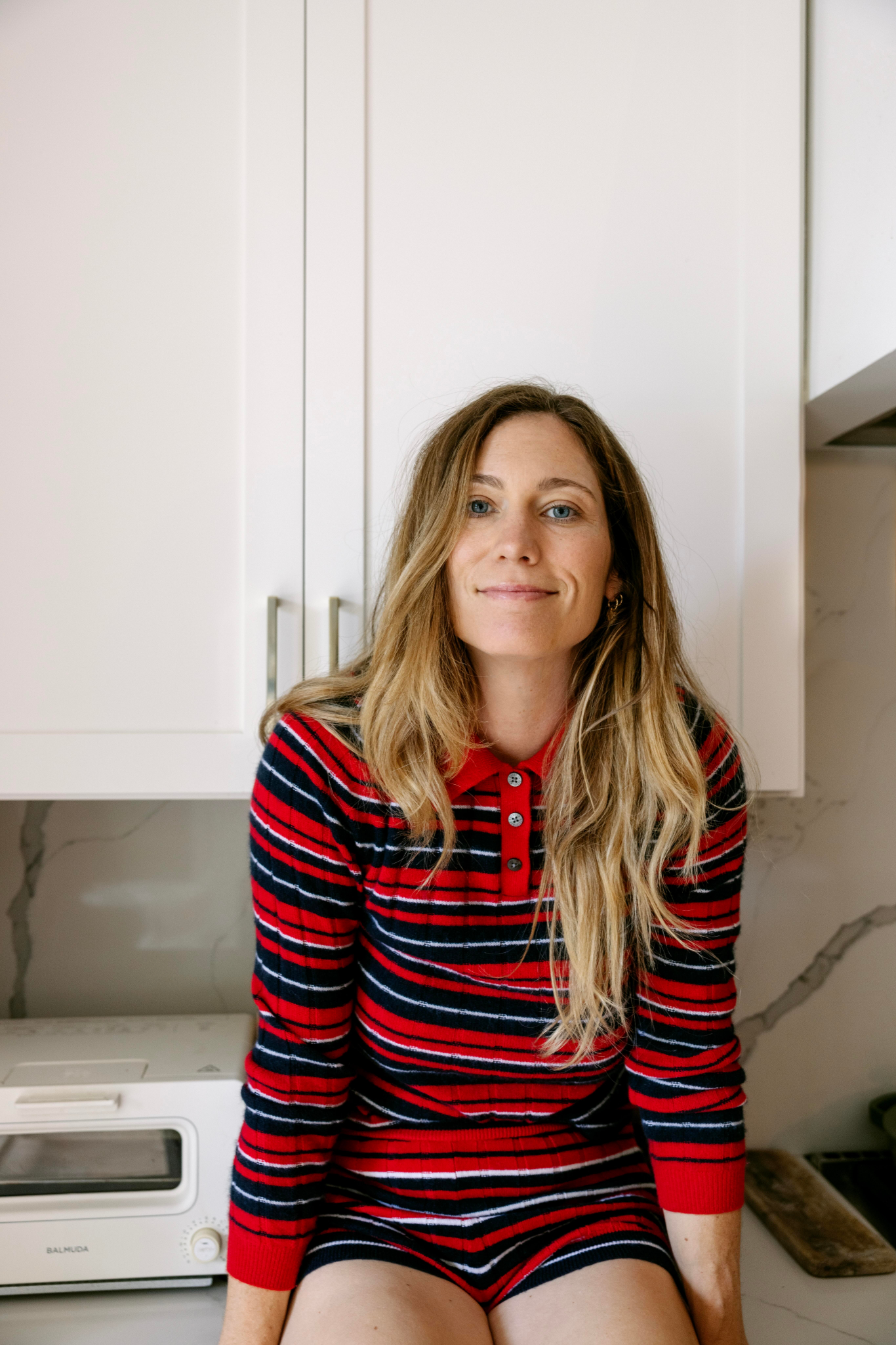 A woman with long blonde hair wearing a red and black striped outfit sits on a kitchen counter in front of white cabinets, smiling slightly at the camera. A toaster oven is visible beside her.