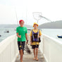 Two boys walk barefoot on a pier, fishing rods and nets in hand, with MIMIKAI Mosquito & Tick Repellent Spray nearby. One sports a red beanie and green shirt, the other a yellow hat and life vest. Boats and hills fill the background.