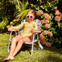A young girl in pink sunglasses and a yellow swimsuit sits on a striped lawn chair in a garden, sticking out her tongue. Surrounded by blooming pink flowers, she enjoys the sun while MIMIKAI Kids Mosquito & Tick Repellent Mist keeps bugs away.