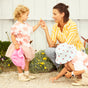 A smiling woman in a yellow striped shirt receives a yellow flower from a young girl, as another child in a cherry sunhat explores nearby—all outdoors with MIMIKAI Kids Mosquito & Tick Repellent Mist among yellow blooms.
