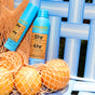 Two blue bottles of MIMIKAI Tick Repellent Mist and three oranges sit in a mesh bag on a white and blue striped chair outdoors in sunlight.