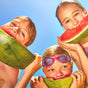 Three children enjoy large watermelon slices outdoors on a sunny day, smiling under the blue sky. One wears purple goggles, and all are protected by MIMIKAI Tick Repellent Mist.