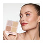 A woman with smooth, radiant skin and peach-toned makeup holds up the Mineral Fusion Highlighter Palette, featuring four neutral shades. She glows with highlighted cheeks and glossy lips, capturing a look of youthful allure.