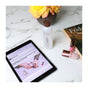 A tablet displaying a beauty article sits next to Mineral Fusion Nail Polish Remover, a bottle of hair product, lipstick, a book, and a vase of yellow flowers on a marble surface.
