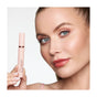 A woman with light skin and blue eyes holds a pink tube of Mineral Fusion So Classy Volume Mascara near her face. She has radiant skin, peach makeup, and slicked-back hair, posing against a plain white background.