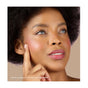 A woman with natural curls and glowing skin touches her cheek, showcasing Mineral Fusions 3-in-1 Color Stick as pink blush and lip tint. She gazes upward confidently against a neutral backdrop.