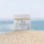 A jar of Sea-el Miracle Kelp Eye Cream rests on sandy ground with a blurred ocean and sky backdrop, emphasizing its beachside origins—your must-have eye cream for reducing dark circles and puffiness.