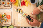 A person slices sushi on the WoodForChef Mizu Minimalist Cutting Board Kit, crafted from 100% FSC-certified maple wood. Sushi, vegetables, soy sauce, ginger, and avocado are nearby. Their hands with rings are visible. Made in Canada.