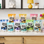 Ten vibrant bags of Molly’s Suds, including Activewear Laundry Powder, sit on a kitchen counter with flowers, a yellow lamp, and stacked jars in the background.
