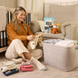 A woman sits on the nursery floor, folding baby clothes and cloth diapers by a laundry basket. Behind her are an armchair, a stuffed animal, and a bag of Molly’s Suds Baby Laundry Detergent Powder.