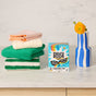 A box of Molly's Suds Color Shield Sheets for Laundry sits on a counter beside folded green, white, and pink towels—ideal for mixed loads—while a blue-and-white striped vase with a yellow flower brightens the background.