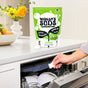 A person puts a Molly’s Suds Dishwasher Detergent Pod into a loaded dishwasher. A bag of Molly’s Suds Dishwasher Detergent Pods sits on the counter with flowers and fruit in the background.