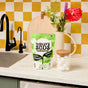 A bag of Molly’s Suds Dishwasher Detergent Pods sits on a white kitchen counter beside a glass jar of pods, with a nearby sink and decorative flowers. A yellow and white checkerboard tile wall adds brightness to the scene.