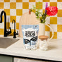 A package of Molly’s Suds Dishwasher Detergent Pods sits on a kitchen counter beside a glass jar of pods, with a sink, flower vase, and yellow checkered backsplash in the background.