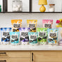 Molly's Suds Laundry Detergent Powder, ideal for sensitive skin, is displayed in assorted packages and scents on a kitchen counter with jars, a yellow lamp, and flowers.