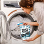 A person in a striped shirt adds Molly's Suds Laundry Detergent Powder to a front-loading washer with white laundry—ideal for sensitive skin.