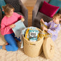 A woman and young girl smile as they fold laundry next to a woven basket filled with colorful towels and a box of Molly’s Suds Laundry Detergent Powder. Pillows and armchairs can be seen in the background.